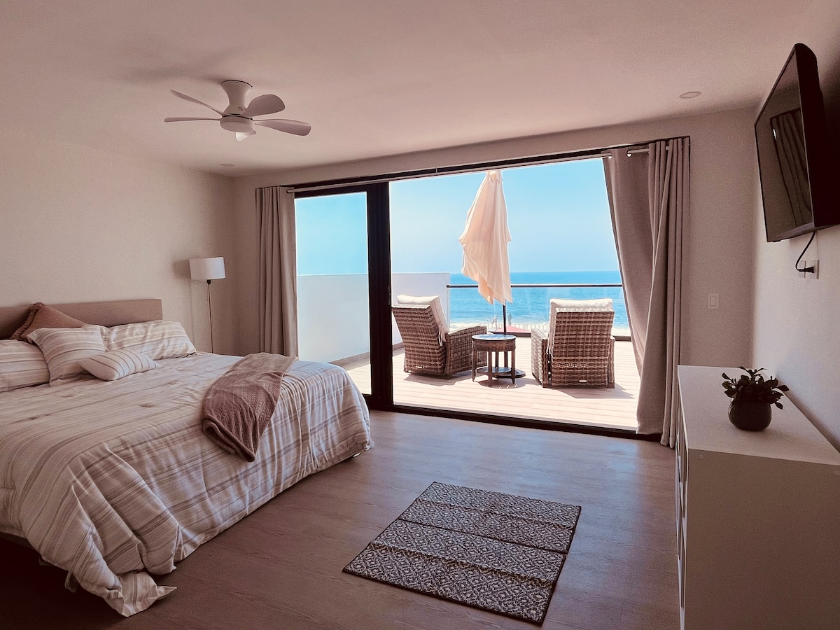 A spacious bedroom features a large bed with light-colored linens, complemented by a ceiling fan. Floor-to-ceiling glass doors provide unobstructed ocean views and lead to a private terrace with two wicker chairs and a small table. A neutral rug adds warmth to the room.