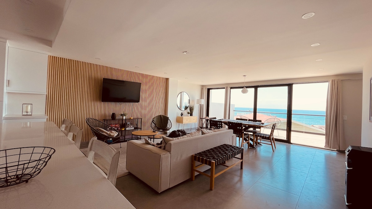 An open living area is highlighted by large glass doors, providing expansive views of the ocean. The space includes a comfortable seating arrangement, a dining area, and a modern kitchen. Natural light illuminates the room, creating a bright and inviting atmosphere.
