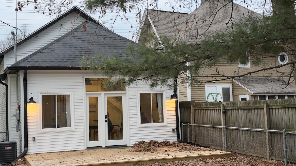 The exterior view of the home showcases a modern design with a white façade and a wooden deck. Large glass doors are flanked by two windows, providing ample natural light. A wooden fence surrounds the backyard, with leaves scattered on the ground, indicating the season.