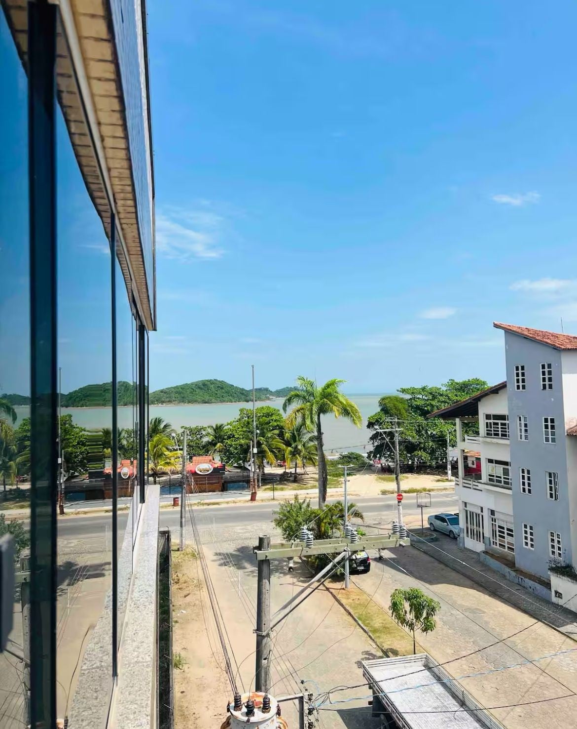 A panoramic view showcases the serene coastline, with green hills visible in the distance and lush palm trees lining the waterfront. The image captures a clear blue sky, creating a bright and open atmosphere. The reflection of the building's windows adds a modern touch to the scene.
