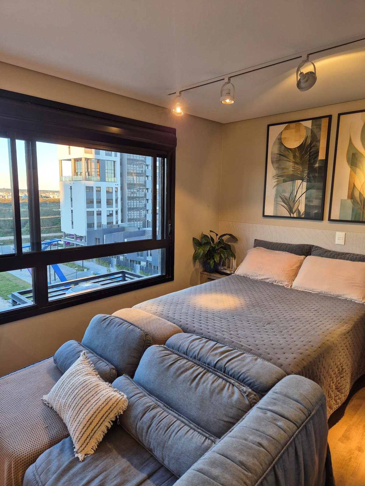 A well-lit bedroom features a spacious bed adorned with soft linens. Two decorative wall art pieces add a touch of style above the bed. A comfortable seating area is positioned beside the window, which offers views of the surrounding landscape.