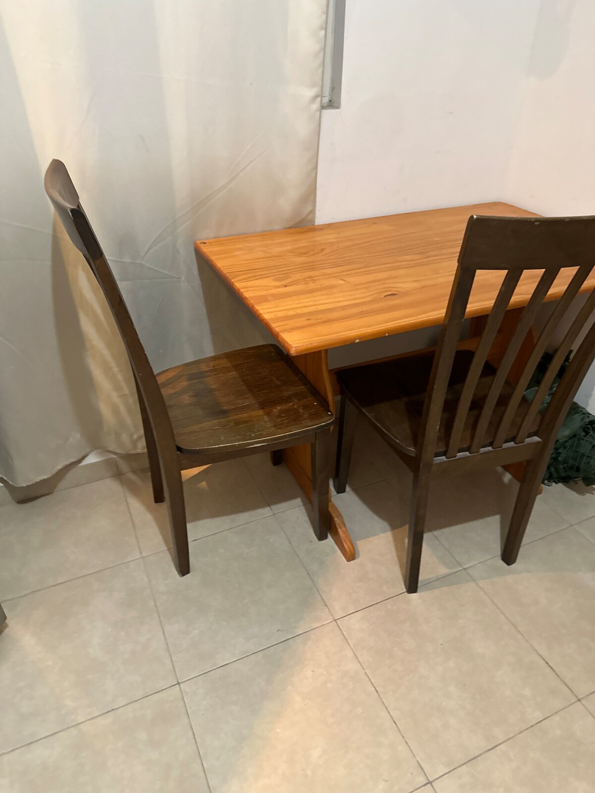 A small, rectangular wooden table is accompanied by two chairs, positioned against a wall. The light wood of the table contrasts with the darker tones of the chairs. A neutral curtain softly drapes in the background, enhancing the simplicity of this dining area.