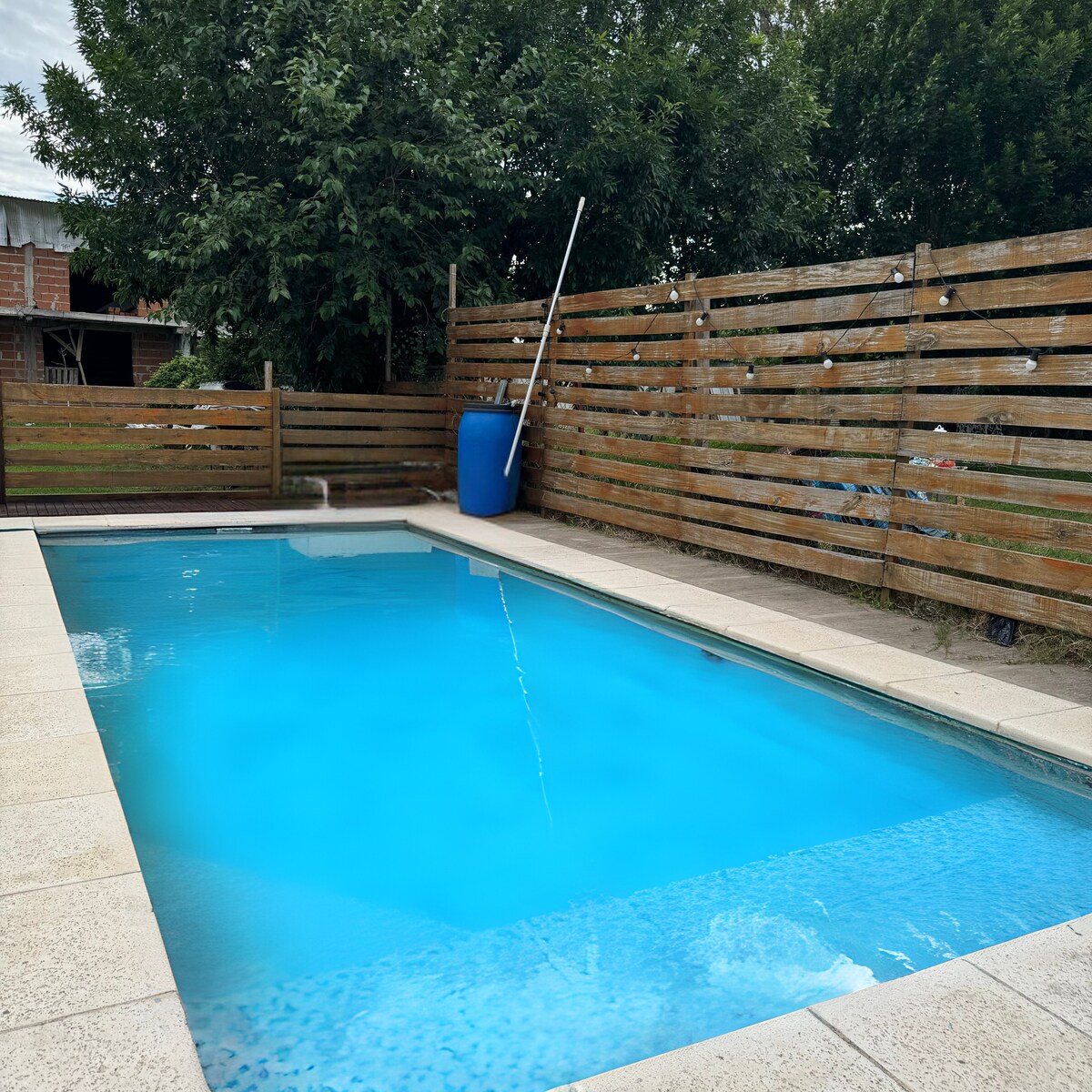 An inviting swimming pool with clear blue water is surrounded by a stone patio. Wooden fencing provides privacy, complemented by a blue barrel positioned beside the pool. Lush greenery is visible in the background, adding to the serene outdoor setting.