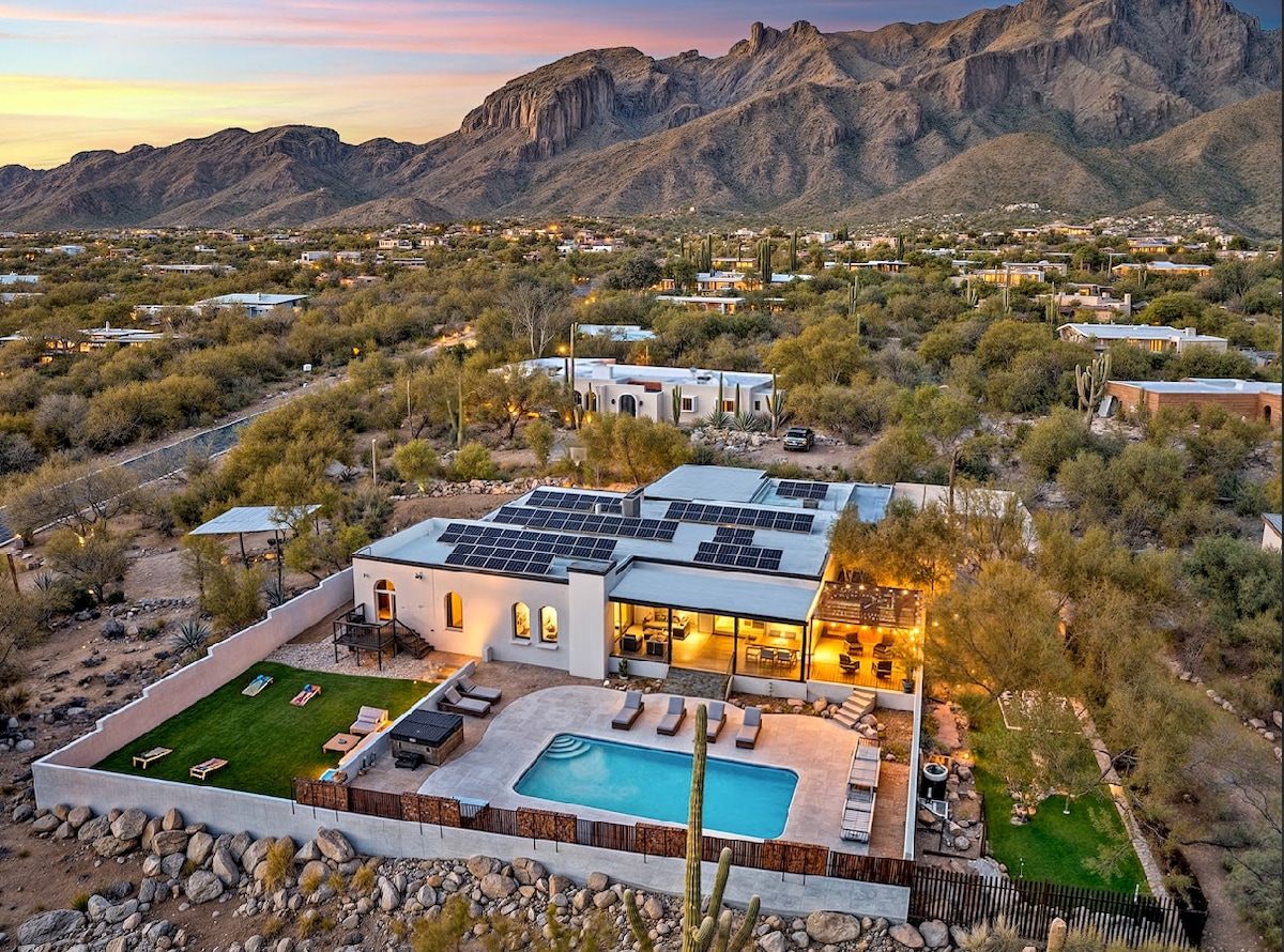 An aerial view showcases the spacious estate nestled in the Catalina Foothills. A heated pool glimmers in the sunlight, surrounded by lounge chairs and a landscaped yard. The backdrop features majestic mountain vistas and serene desert landscaping, creating a peaceful retreat atmosphere.