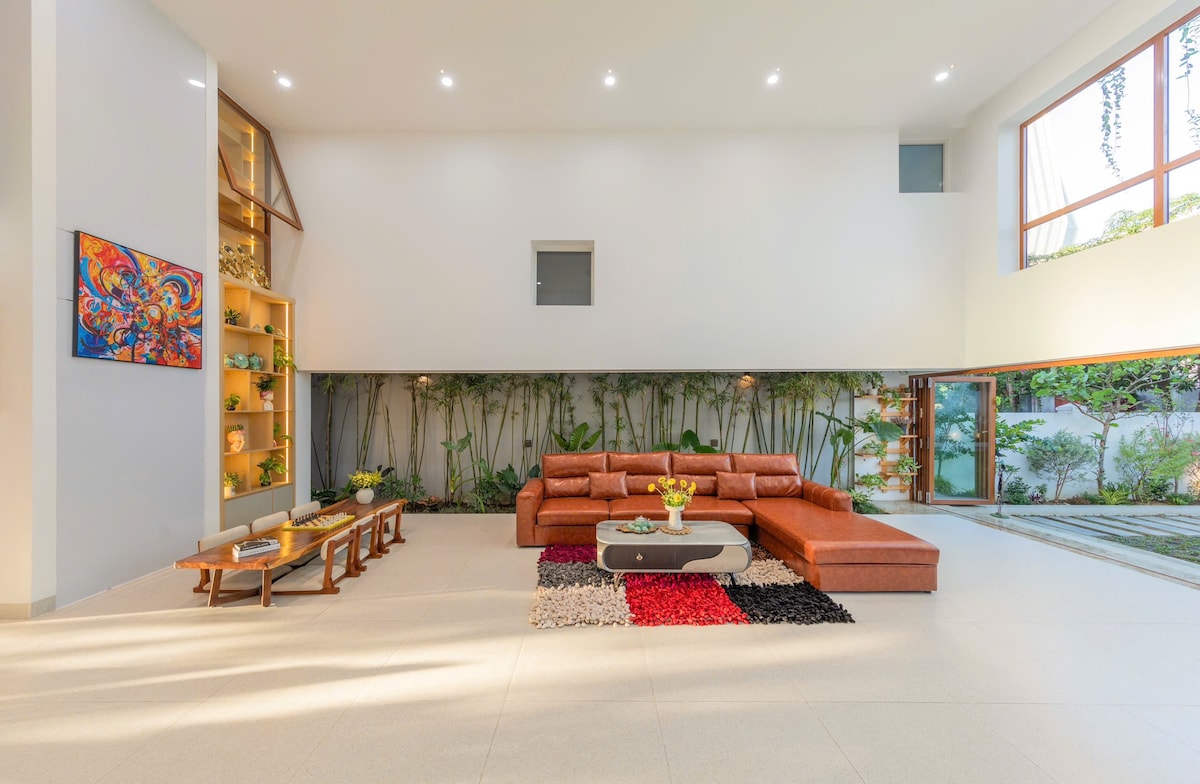 A spacious living room features an L-shaped leather sofa arranged around a low coffee table. Natural light floods through large windows, illuminating the neutral-toned walls and floor. Decorative shelves are displayed with greenery and art, while a colorful area rug adds a touch of vibrancy.