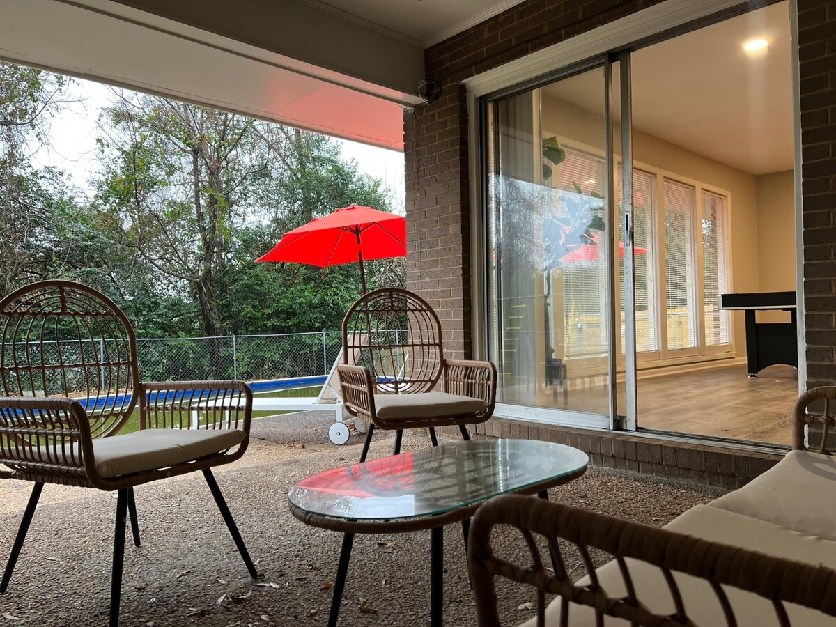 An outdoor patio area features two rattan chairs and a glass table, inviting relaxation. Large sliding doors open to a bright interior space, while bright red umbrellas provide shade against the backdrop of a lush, green backyard, which includes a visible pool.