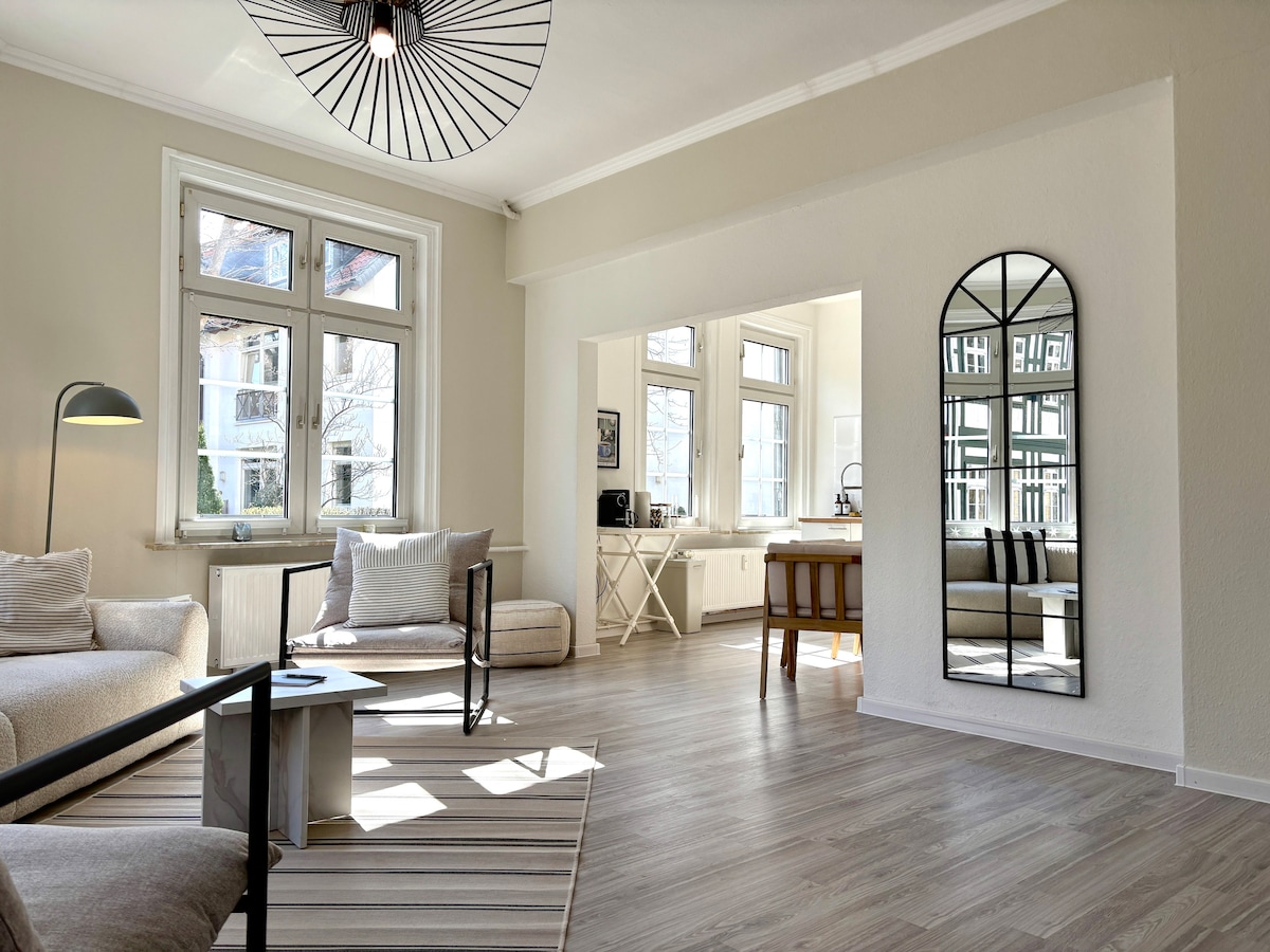 The spacious living area features large windows allowing natural light to fill the room. A comfortable sofa and chair are positioned on a light-striped rug, while a modern coffee table sits in the center. A mirror and a dining area are also visible, enhancing the room's openness.