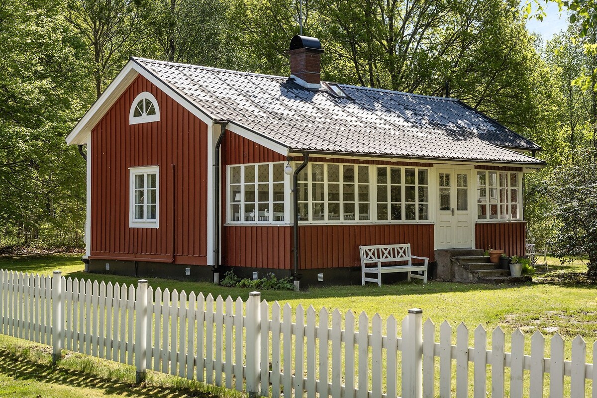 A charming red wooden cottage is surrounded by lush greenery. Large windows allow for ample natural light, while a welcoming porch features a white bench. A white picket fence encloses the property, enhancing the inviting rural atmosphere.