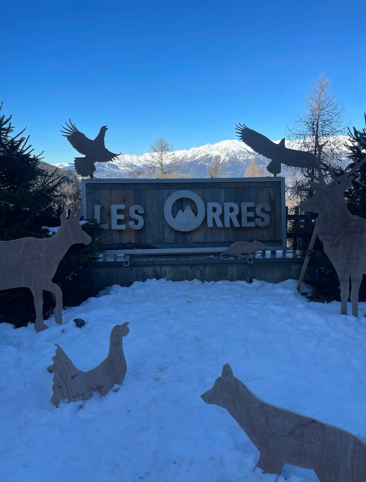 A sign displays 'Les Orres', surrounded by wooden animal silhouettes. Snow blankets the ground, with a backdrop of distant snow-capped mountains and clear blue skies, suggesting a serene winter landscape.