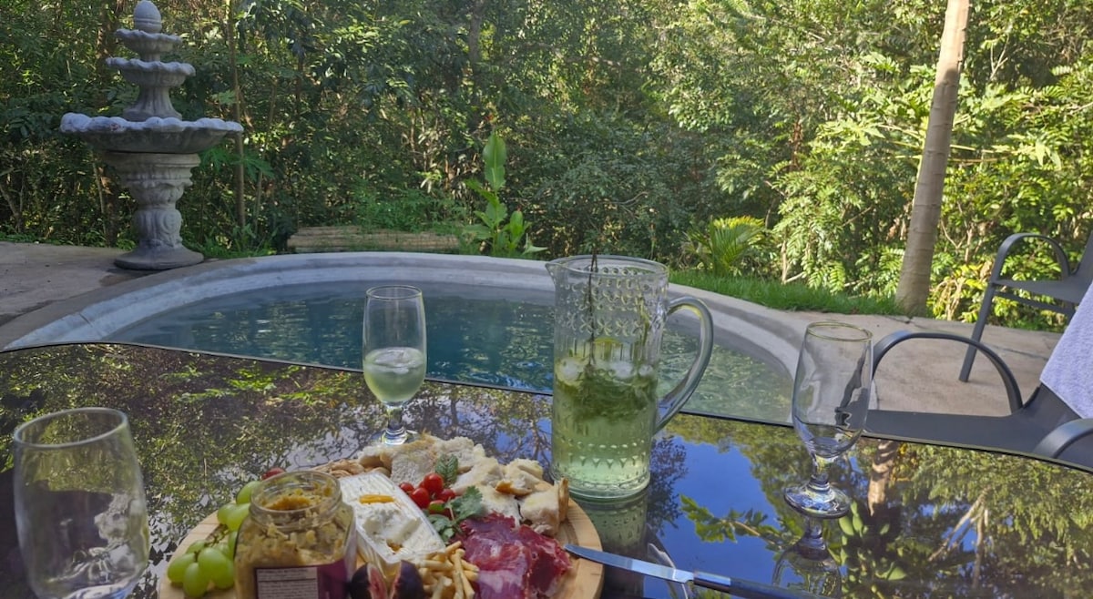 A glass table displays an assortment of snacks, including cheese and fruits, beside two glasses of beverages. In the background, a serene swimming pool reflects lush greenery, with a fountain nearby adding a sense of tranquility to the setting.