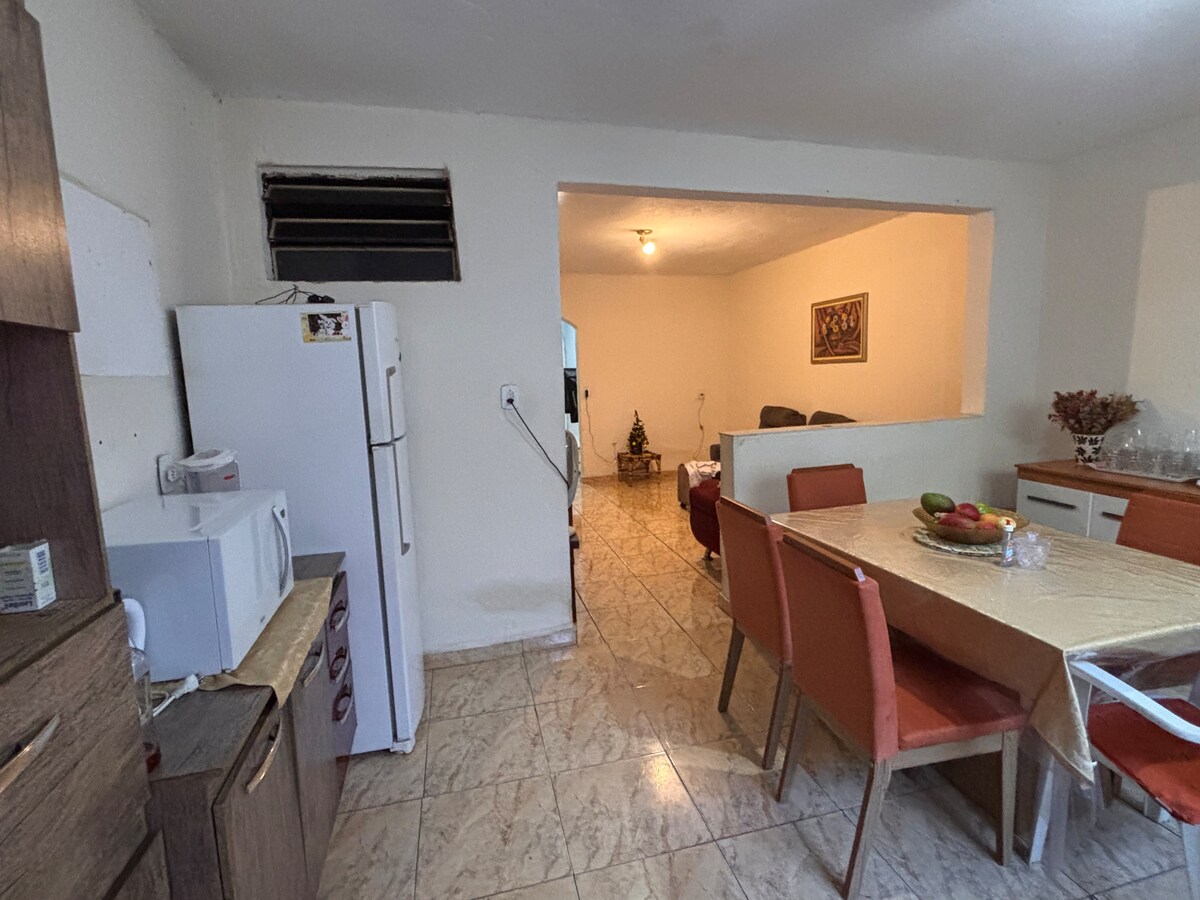 The open-concept kitchen and dining area features a wooden dining table with orange chairs. A microwave and refrigerator are visible, while a living area can be glimpsed in the background. Neutral-colored walls complement the tile flooring throughout the space.