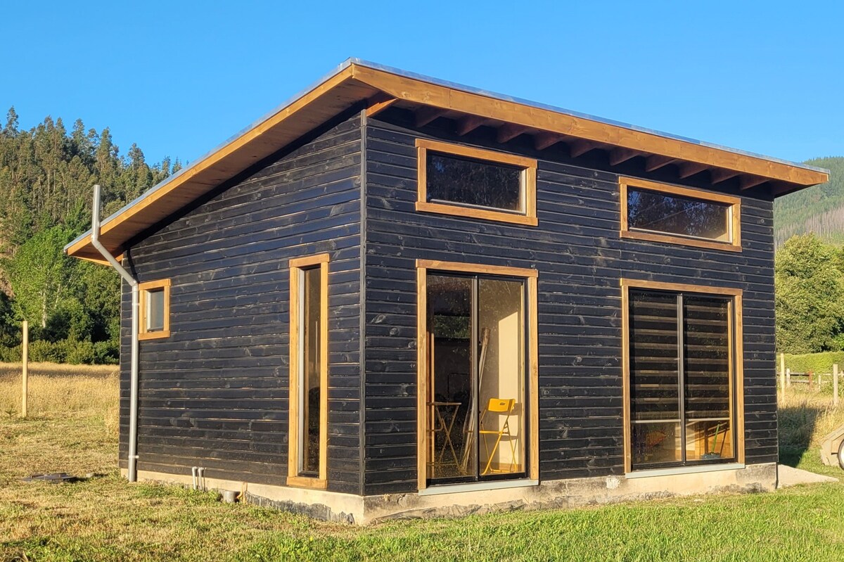 A modern cabin features a sleek black exterior with warm wooden accents, including large windows that invite natural light. The surrounding landscape opens up to grassy fields and distant trees, offering a serene backdrop and a connection to nature.