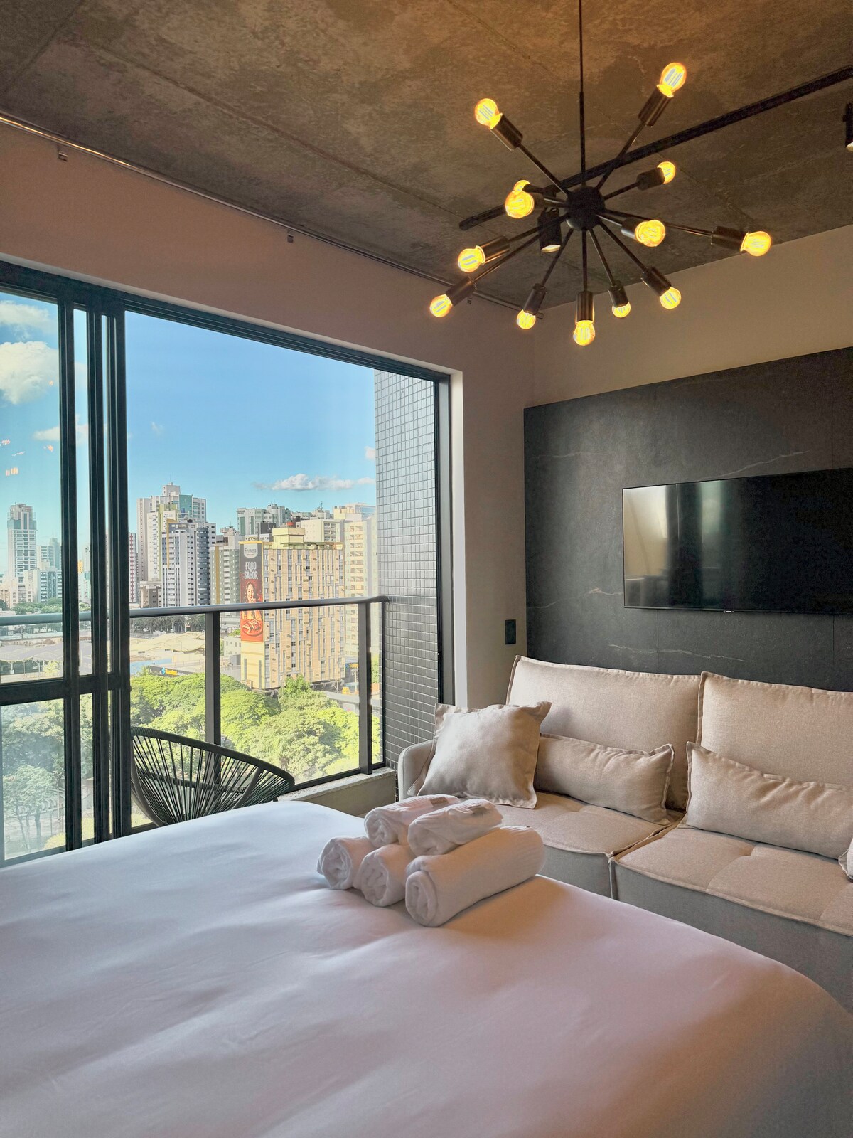 A bright room features a spacious bed adorned with neatly arranged towels. A comfortable sofa offers a cozy seating area, positioned near large glass doors that provide a view of the city skyline, with natural light flooding the space.