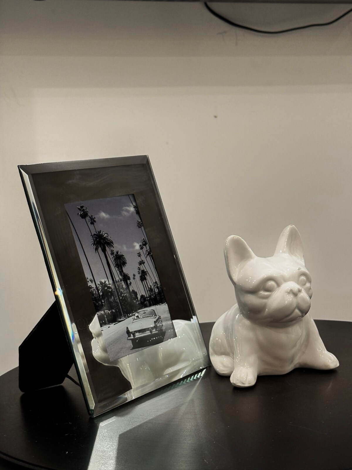A decorative table features a sleek, mirrored picture frame displaying a black-and-white photo with palm trees and a classic car. Beside it, a white ceramic figurine of a French bulldog sits, adding a charming touch to the arrangement.