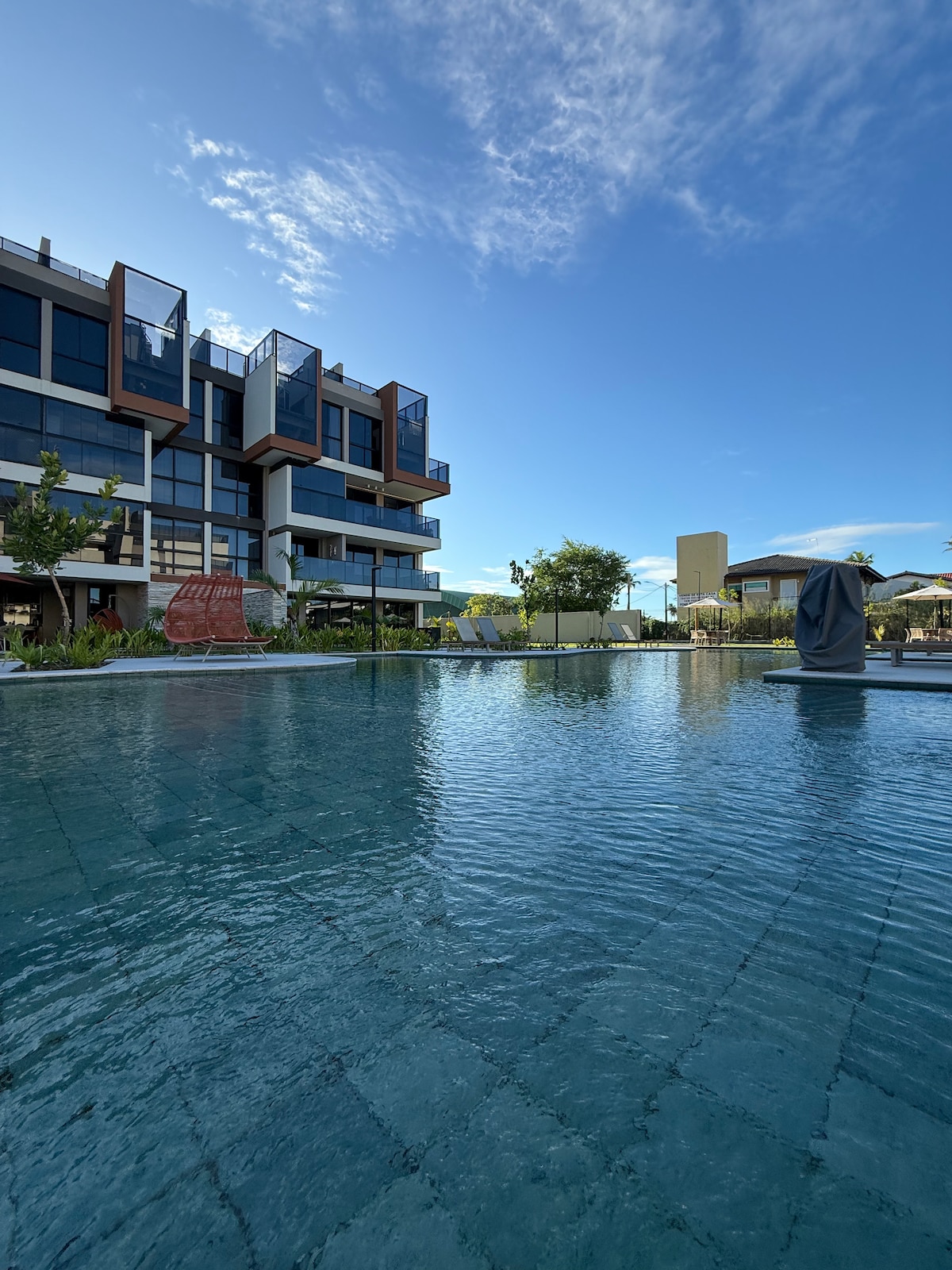 A modern building with large glass windows is visible alongside a tranquil swimming pool. Lush greenery surrounds the area, creating a serene environment. The clear blue sky reflects on the water's surface, enhancing the inviting atmosphere of the outdoor space.