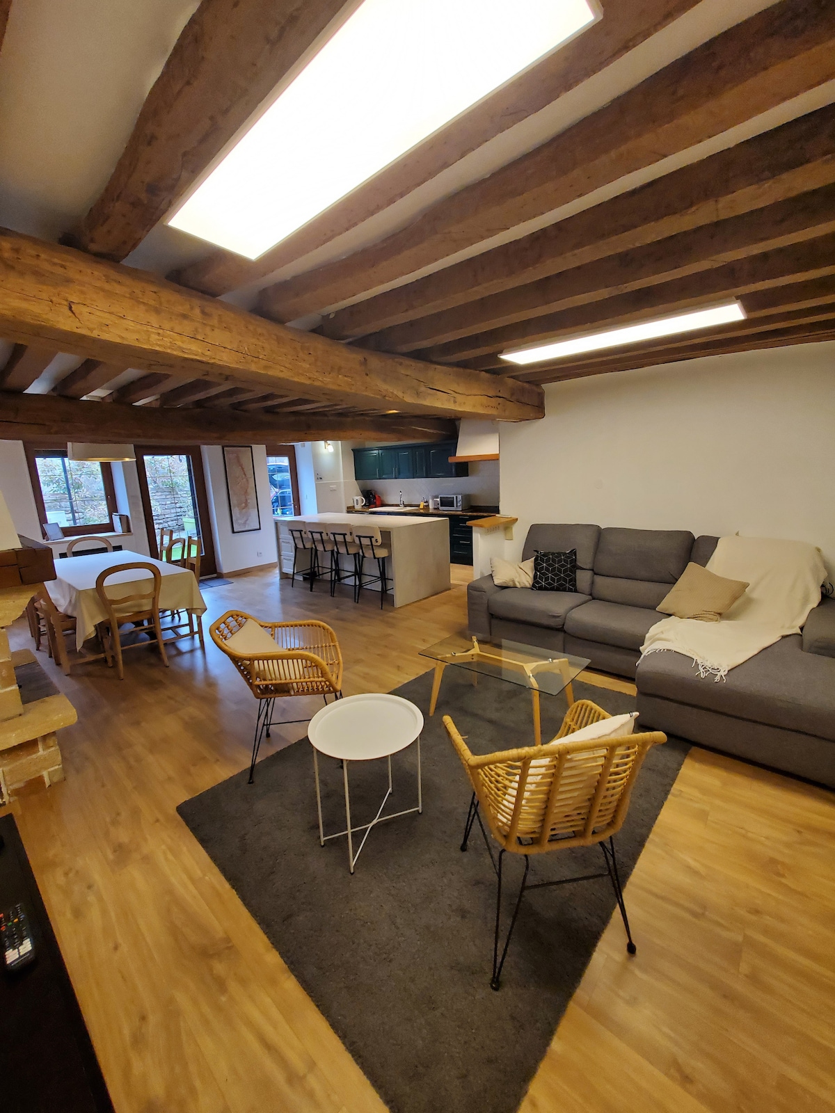 A spacious living area features wooden beams along the ceiling. A comfortable gray sofa is positioned across from two rattan chairs, surrounding a small white table on a soft rug. Natural light pours in through large windows, providing views of the outdoors and an inviting atmosphere.