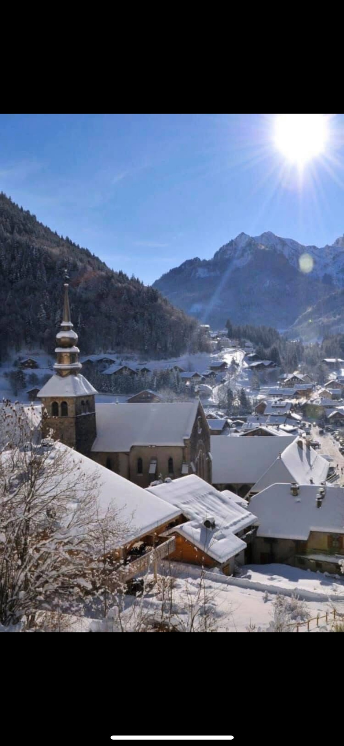 A winter landscape reveals a charming village nestled in snow-covered mountains. The image features the spire of a church rising above the rooftops, all blanketed in white snow. Sunlight glistens off the snowy terrain, adding a serene quality to the scene.