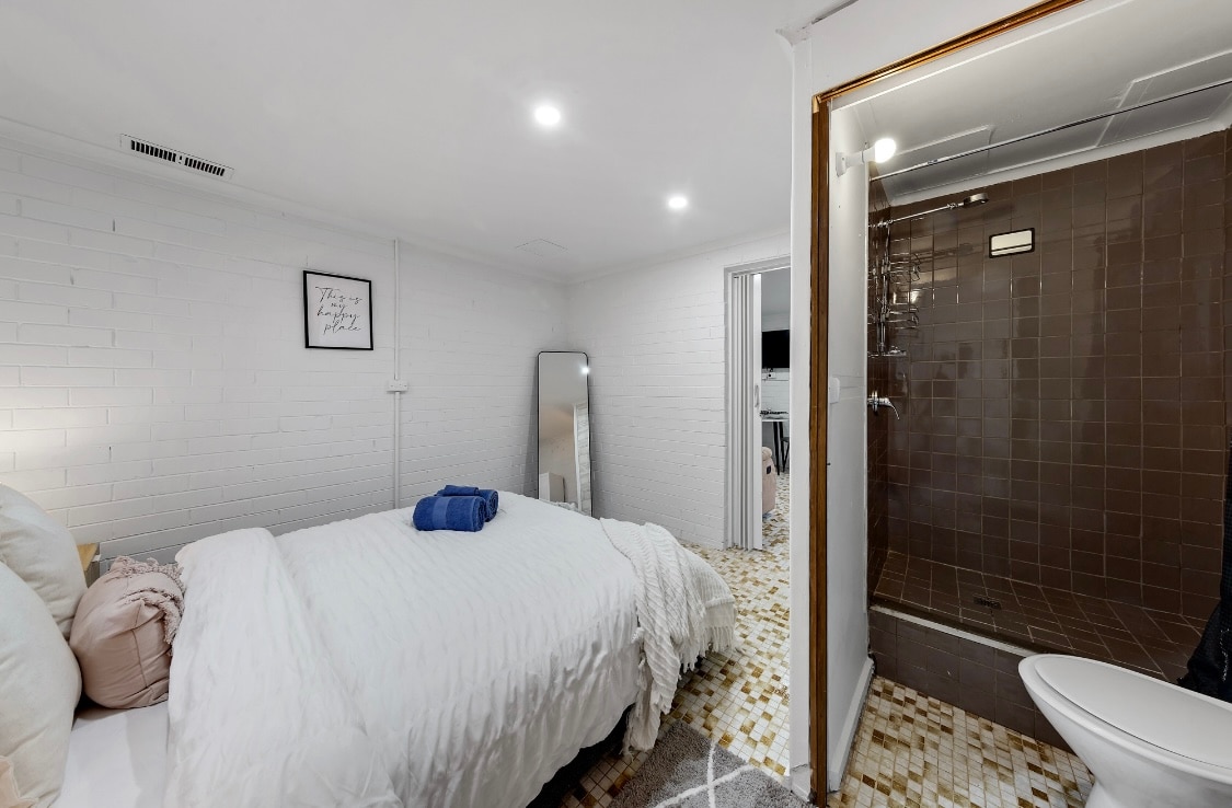A cozy sleeping area is presented, featuring a spacious bed with soft linens and complimentary towels. A full-length mirror reflects the room, while an attached bathroom offers a medium-sized shower and modern fixtures. Light illuminates the white brick walls, enhancing the inviting ambiance.