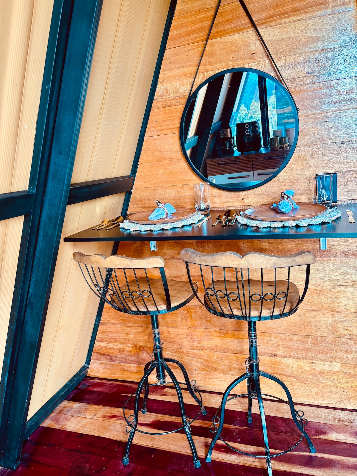 A compact dining area is integrated into the wooden wall, featuring two high-back stools and a circular mirror. The table is set with dining ware, and a small decorative item is placed on the table, enhancing the cozy ambiance of the space.