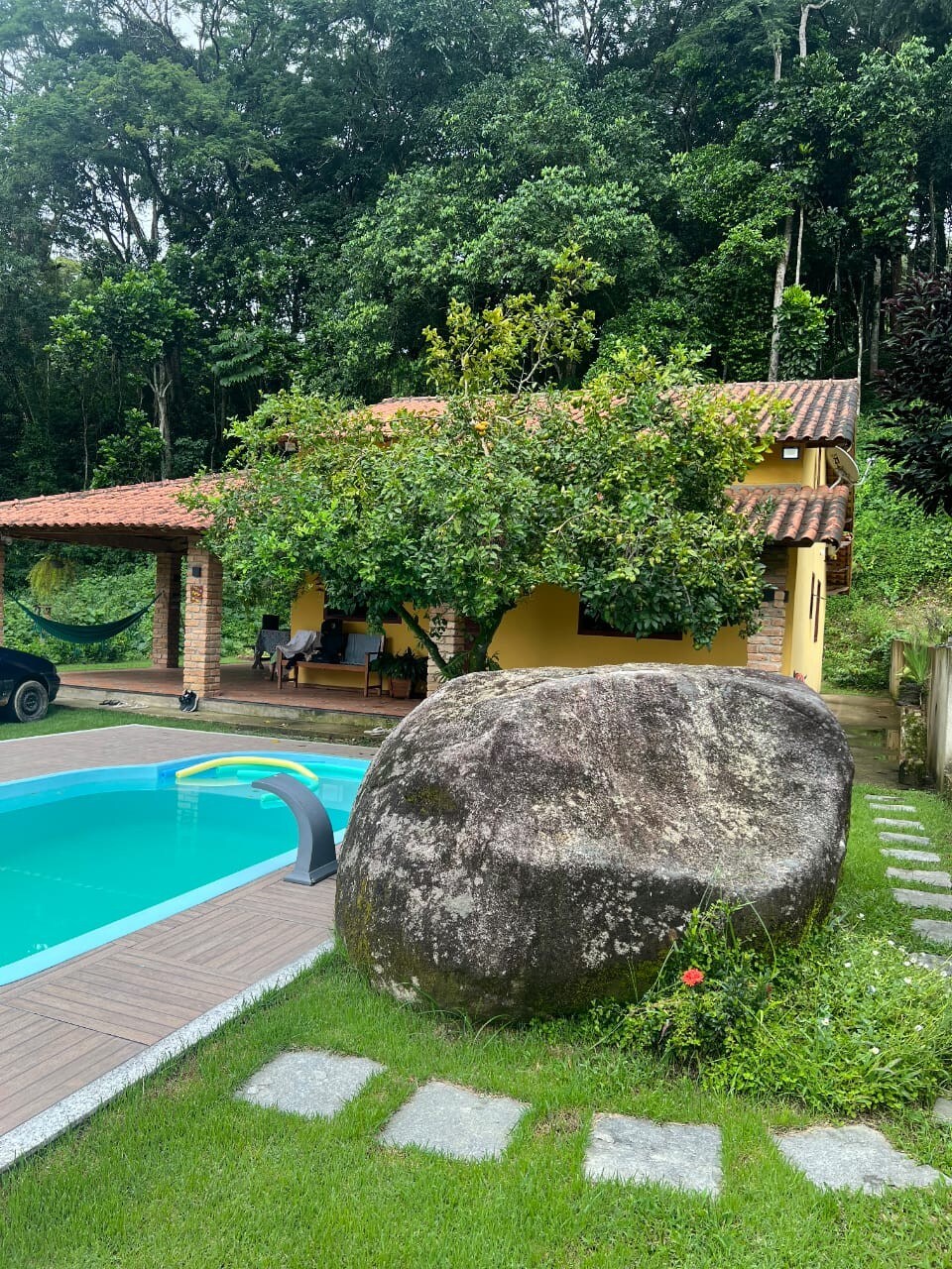 A yellow house set amidst lush green foliage features a rustic roof. A large rock is positioned near a turquoise pool, with wooden decking surrounding the area. A hammock hangs from a tree, offering a spot for relaxation in this tranquil outdoor setting.