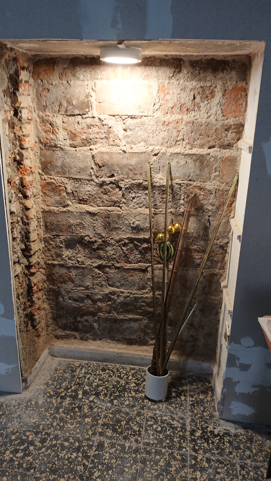 An alcove is highlighted by a soft overhead light, showcasing a textured brick wall. In the corner, a slender vase holds tall, minimalist branches, bringing a touch of nature to the space. The floor features a subtle patterned surface, enhancing the area’s warmth.