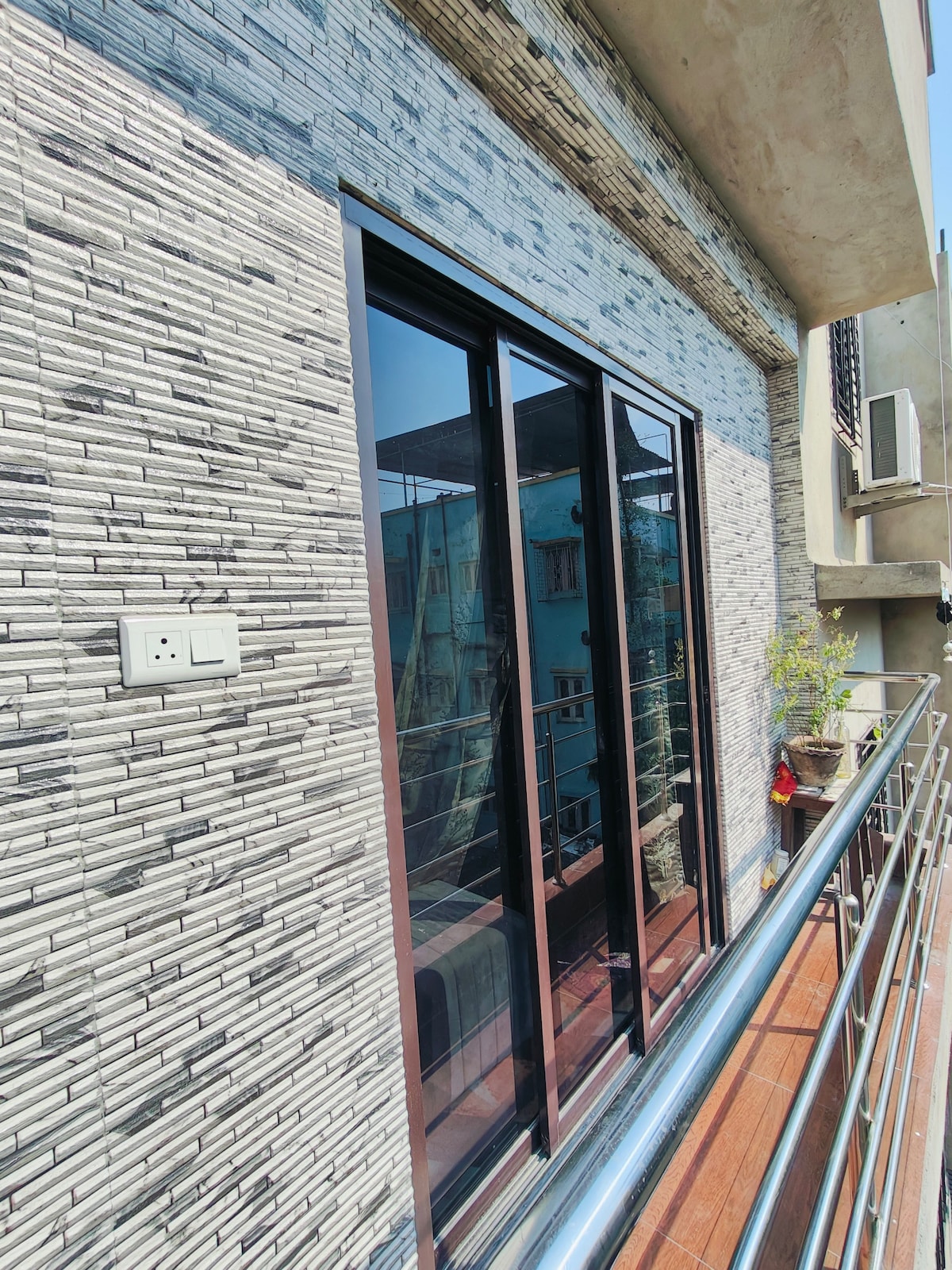 A balcony is depicted with sleek sliding glass doors, offering an unobstructed view of the surroundings. The modern stone-patterned exterior creates a textured backdrop, while a small potted plant adds a touch of greenery. A stainless steel railing provides safety and elegance.