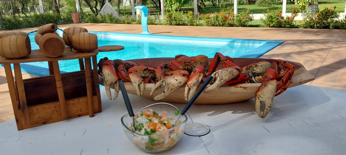 A large, wooden bowl filled with freshly prepared crabs is positioned next to a clear blue pool. A glass bowl of colorful salad sits nearby on a white tablecloth. Lush greenery and blooming flowers surround the pool area, enhancing the inviting setting.