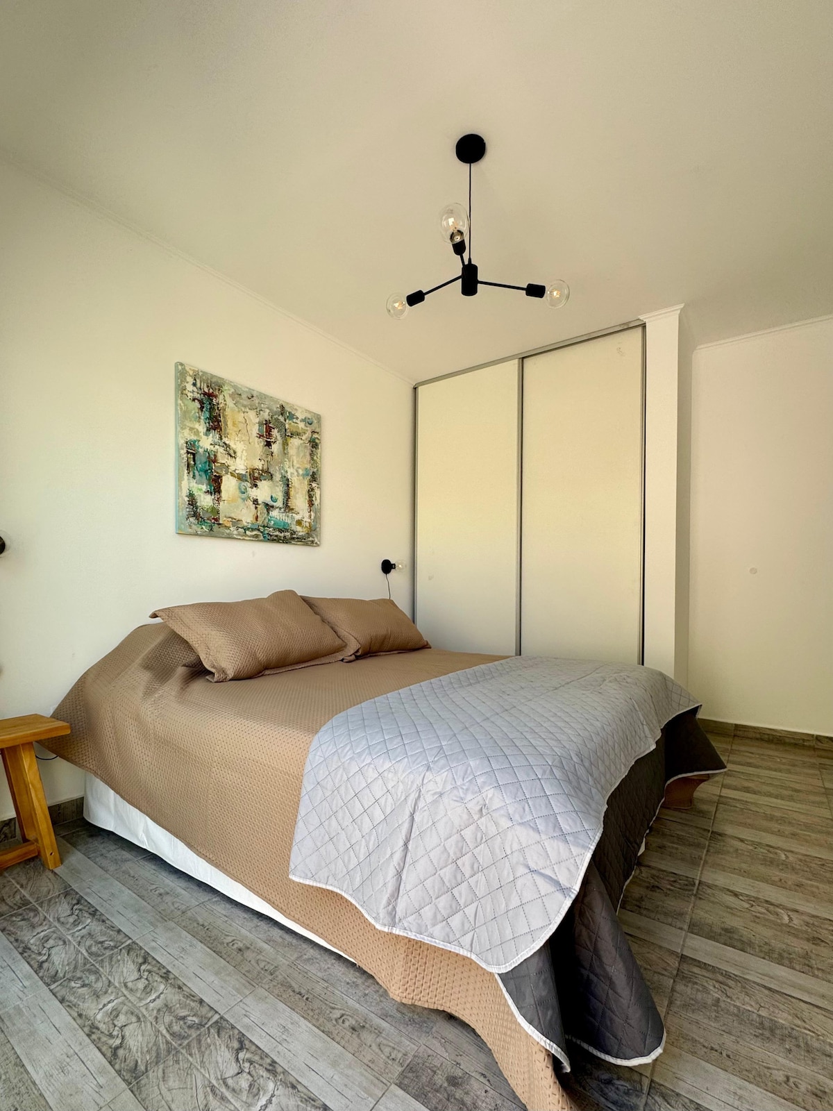 A cozy bedroom features a spacious bed with neutral bedding, complemented by a textured throw. A piece of wall art adds color to the light-toned walls, while a modern light fixture provides overhead illumination. A wardrobe is subtly integrated into the room's design.