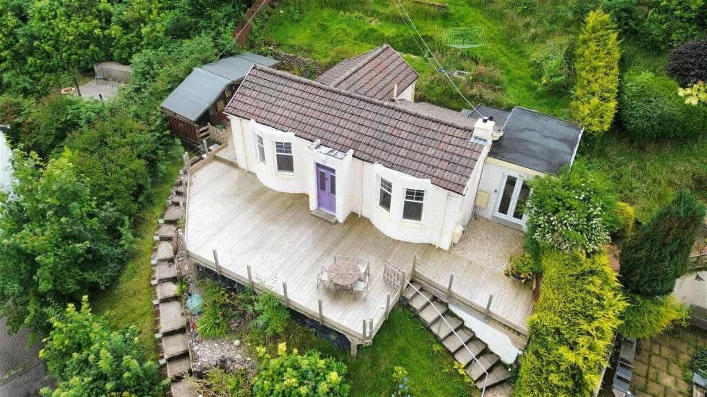 An aerial view captures the unique shape of the art deco home surrounded by abundant greenery. A spacious decked terrace features a small seating area with a table and chairs, accessible via a landscaped pathway leading to the surrounding garden.