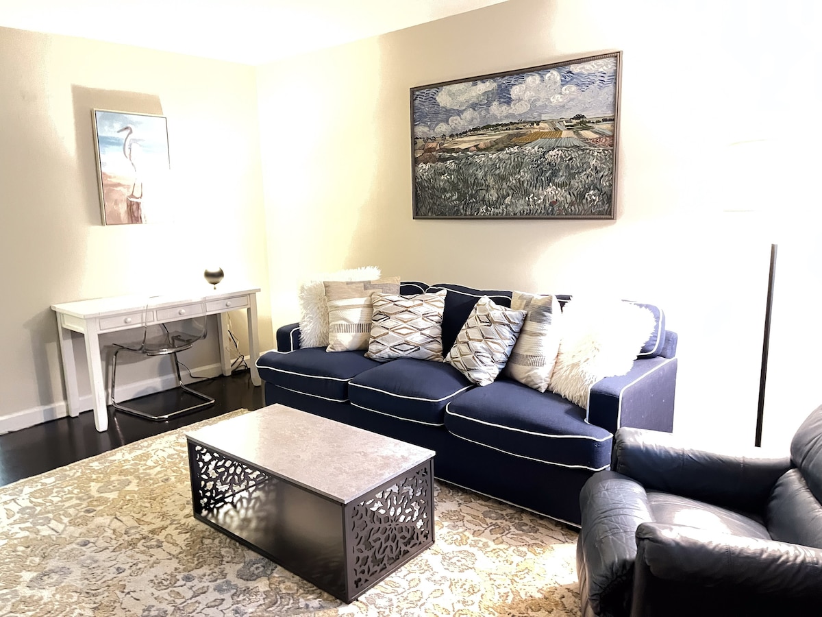 A cozy living room is equipped with a navy blue sofa adorned with decorative pillows. A light-colored coffee table sits on a patterned rug, while a glass writing desk is positioned against the wall, complemented by an artwork above the sofa depicting a serene landscape.