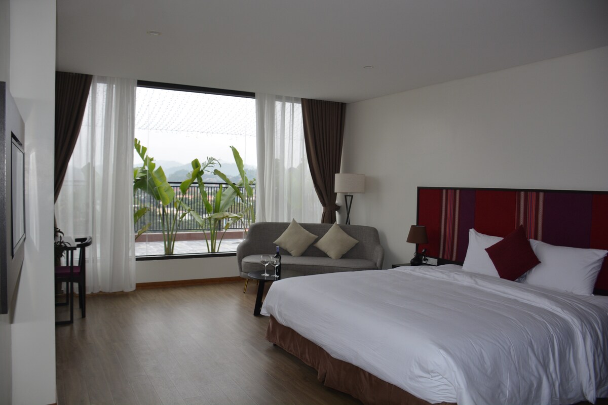 A spacious bedroom features a large bed with crisp white linens and decorative cushions. A comfortable sofa is positioned by a large window, showcasing views of greenery outside. Soft natural light filters through sheer curtains, complementing the neutral tones of the room.