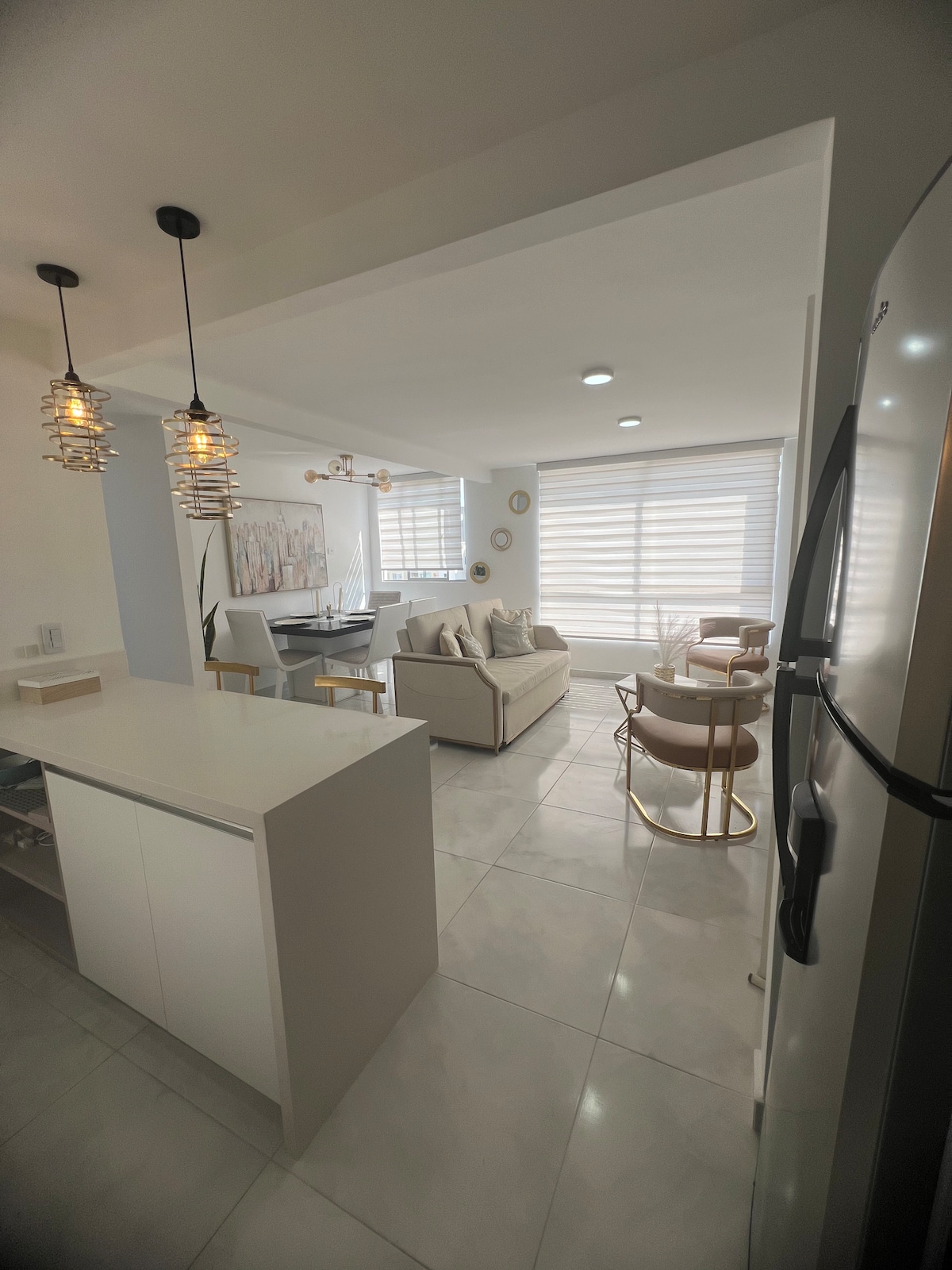 An open-plan kitchen and living area displays modern design with white cabinetry and polished tile flooring. A dining table for four and a cozy seating arrangement are visible, illuminated by natural light through window shades. Pendant lighting adds a warm touch to the space.