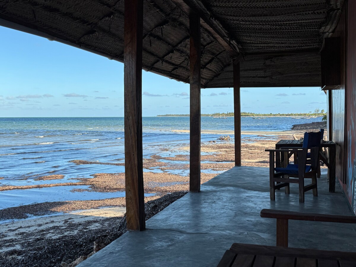 A covered patio offers a view of the serene beach and calm sea. Wooden chairs are positioned around a table, inviting relaxation. The coastline features gentle waves lapping against the shore, with a stretch of sand and rocks visible at low tide under a clear blue sky.