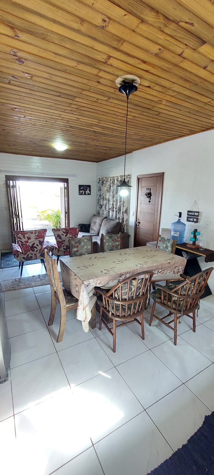 A dining area is presented, featuring a wooden table surrounded by six chairs. A cozy seating space with a patterned chair is visible in the background. Natural light enters through an open window, highlighting the room's warm wooden ceiling and tiled floor.