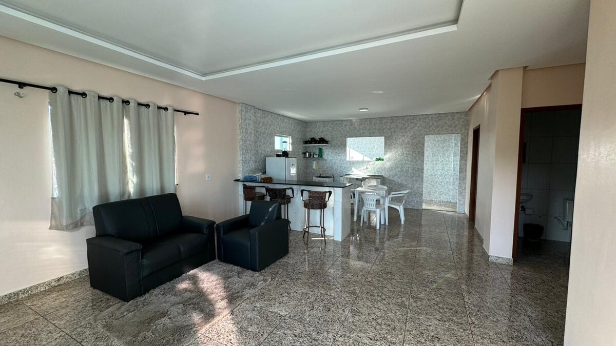 A spacious living area features a black sofa and armchair, accompanied by a dining set for four. The kitchen is open, showcasing modern fixtures and ample counter space. Natural light floods the space through large windows and a partially visible entrance.