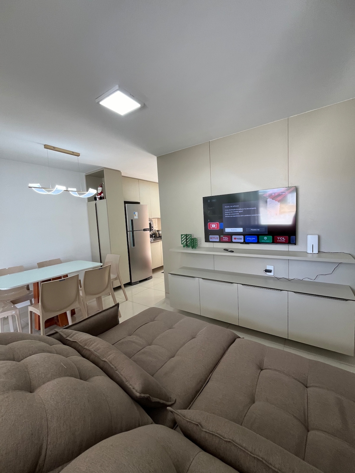 The living area features a comfortable brown retractable sofa facing a wall-mounted smart TV. A dining table with white surface and light-colored chairs is visible in the background, with a modern kitchen accessible through an open layout.