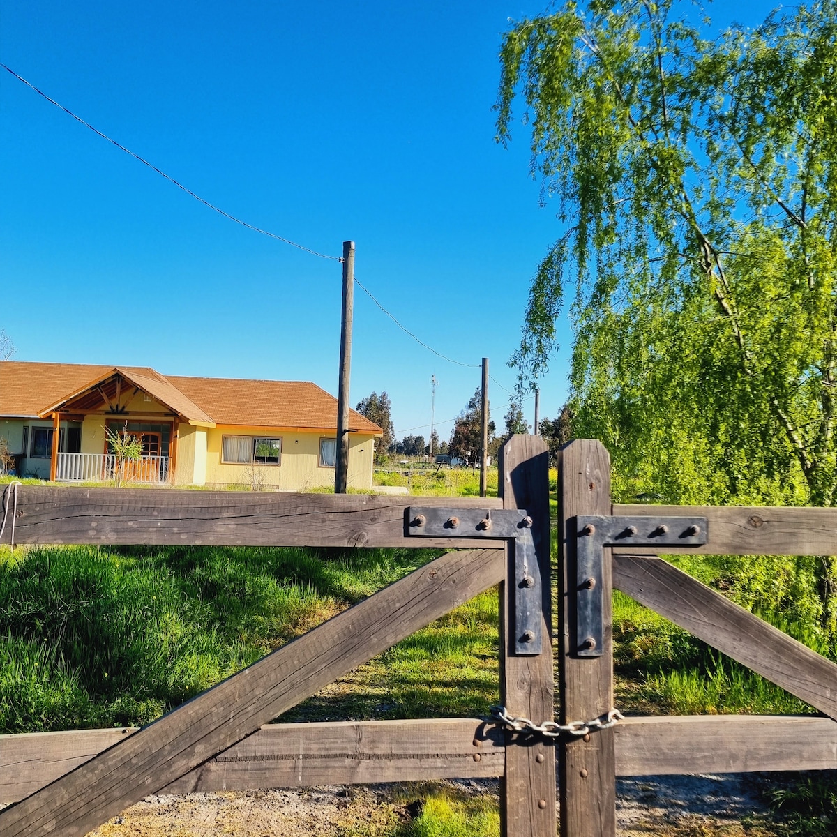 A rustic wooden gate is captured in the foreground, adorned with metal hardware. A yellow house with a brown roof is visible in the background, surrounded by lush greenery and clear blue skies.