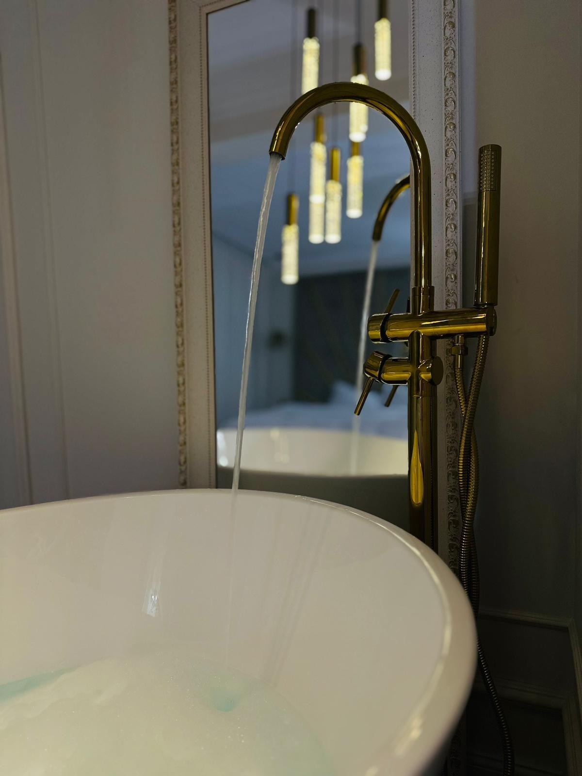A sleek freestanding bathtub is positioned beside a mirror, with water flowing gracefully from a gold faucet. The subtle glow of pendant lights above enhances the elegance of the space, while the presence of airy bubbles in the tub adds to the relaxing ambiance.