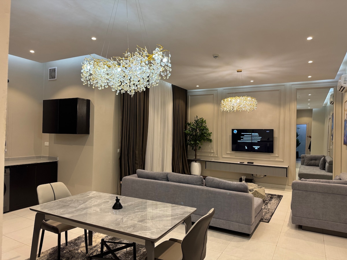 An open living area features stylish furnishings, including two gray sofas and a marble dining table. A television is mounted on the wall opposite the seating area. Soft lighting is provided by decorative chandeliers, while large windows are dressed in dark curtains, allowing for ample light.