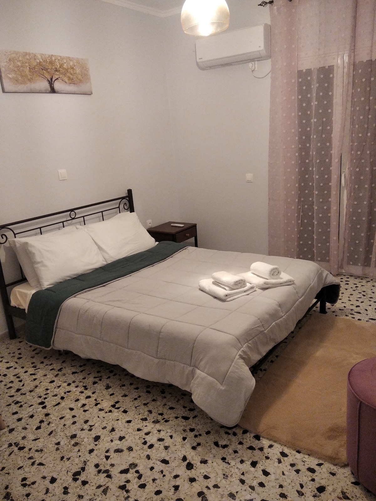 A comfortable sleeping area features a bed with a light quilt and several neatly arranged towels. Natural light enters through a sheer curtain covering a window. A nightstand sits beside the bed, and a purple ottoman is positioned in the corner, enhancing the room's functionality.