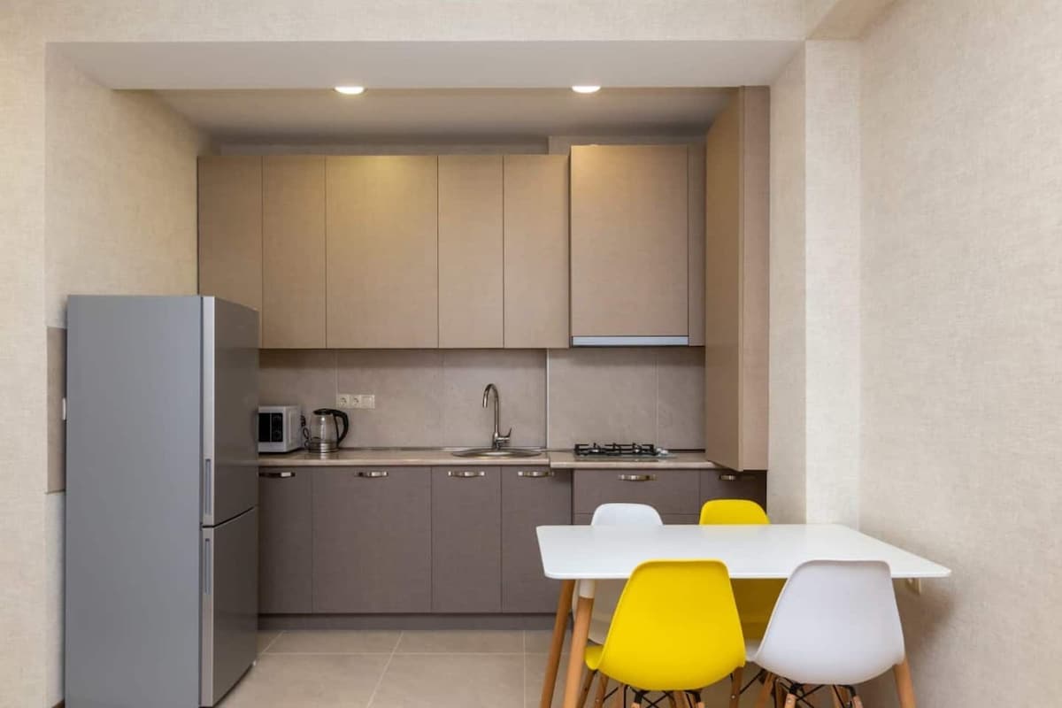 A modern kitchen is presented, featuring sleek cabinetry and gray tones. Stainless steel appliances, including a refrigerator and microwave, are visible. A small table with white and yellow chairs is placed near a window, creating a functional dining area.