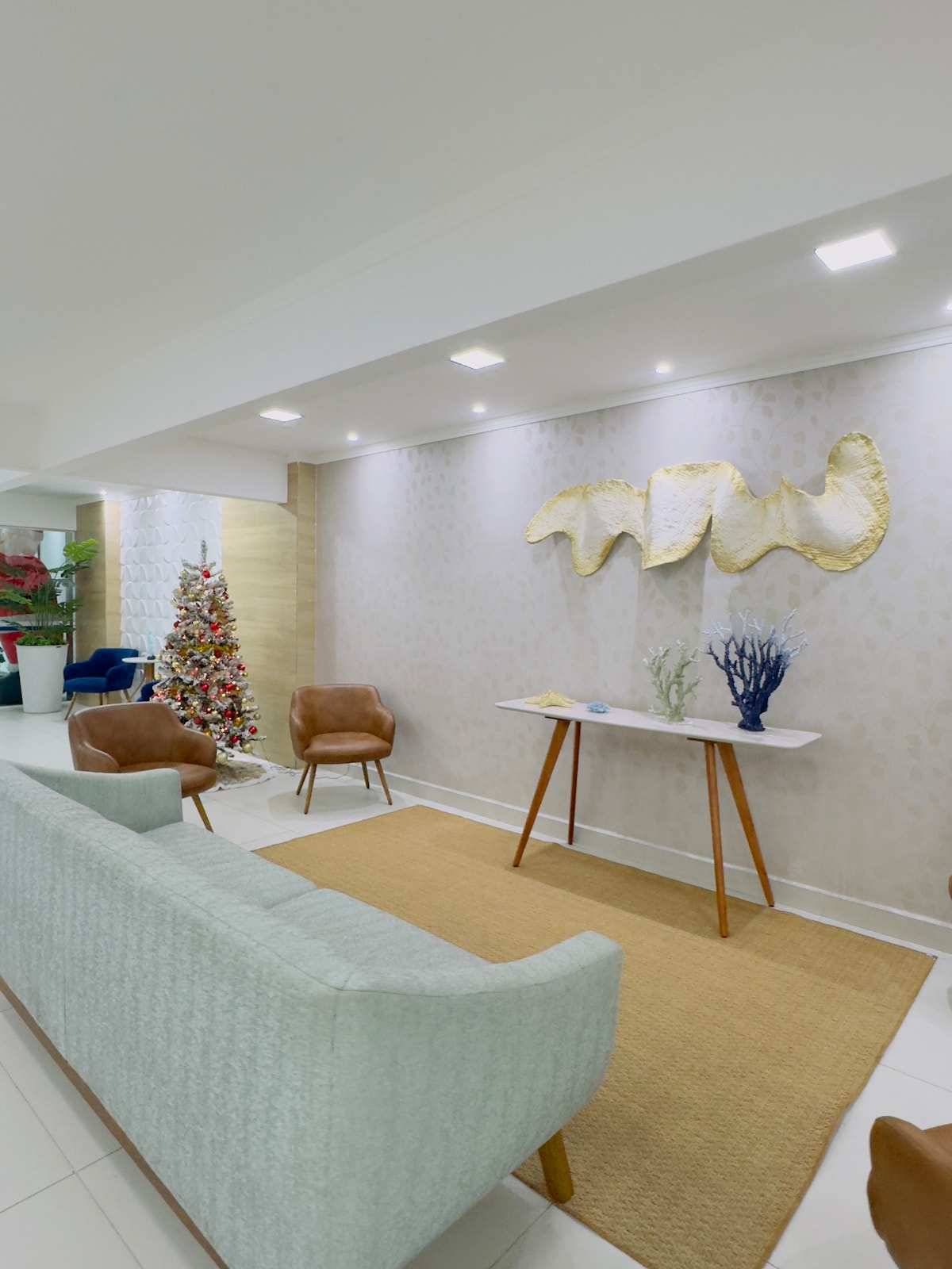 A modern seating area features a soft, light-colored sofa facing two brown chairs. A simple wooden table is positioned in front of the sofa, adorned with decorative elements. The wall behind displays an abstract art piece, illuminated by warm lighting, enhancing the welcoming ambiance.