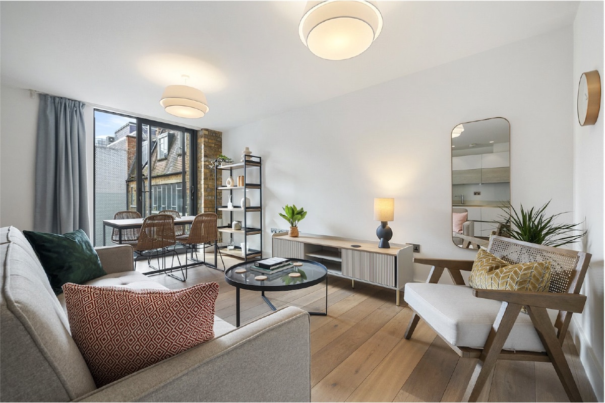 An open-plan living area showcases an inviting seating arrangement with a light gray sofa and a wooden accent chair. Natural light filters through floor-to-ceiling windows, illuminating a round coffee table with decorative items. A shelf holds plants and creates a calming atmosphere.