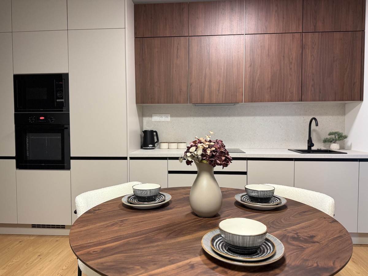 A modern kitchen is highlighted, featuring sleek cabinetry in neutral tones. A round wooden dining table is set with tasteful plates and bowls. A small vase with dried flowers is centrally placed, and appliances are subtly integrated into the kitchen design.