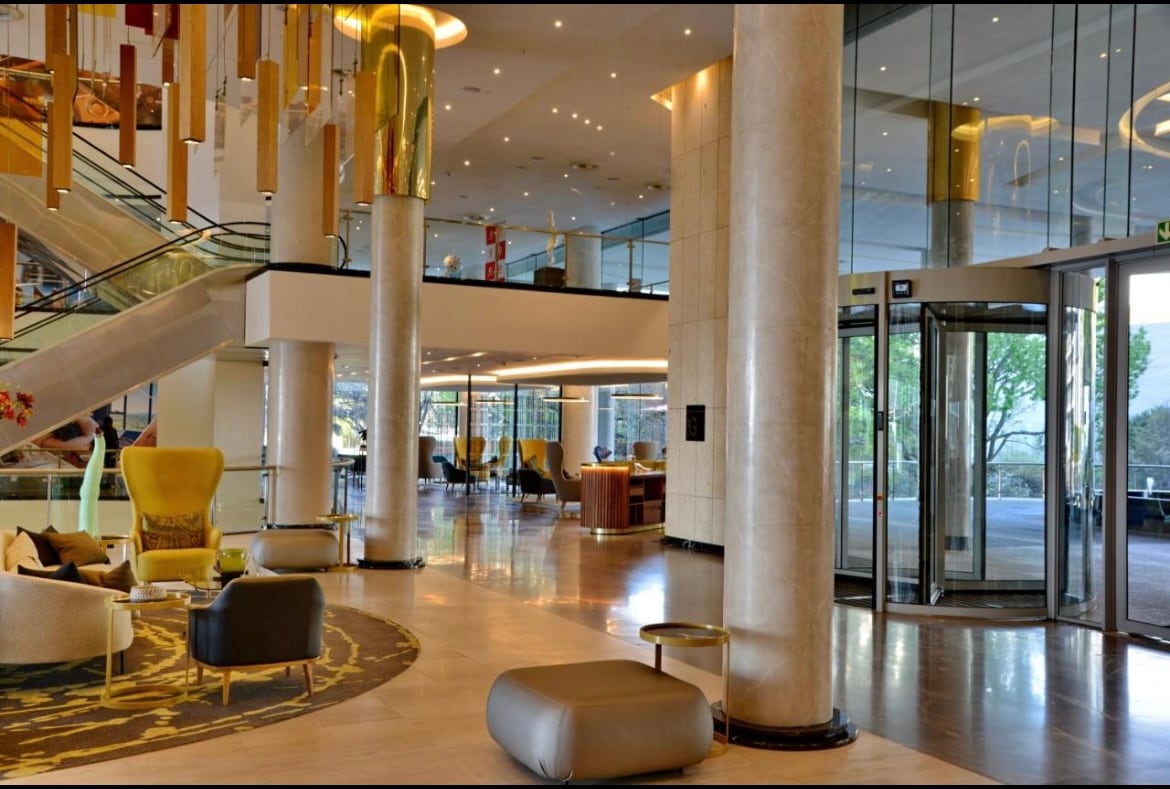 The spacious lobby area features tall glass windows allowing natural light to brighten the space. Elegant pillars and modern furnishings, including a variety of seating options, create an open atmosphere. A subtle color palette complements the stylish decor, enhancing the overall aesthetic.