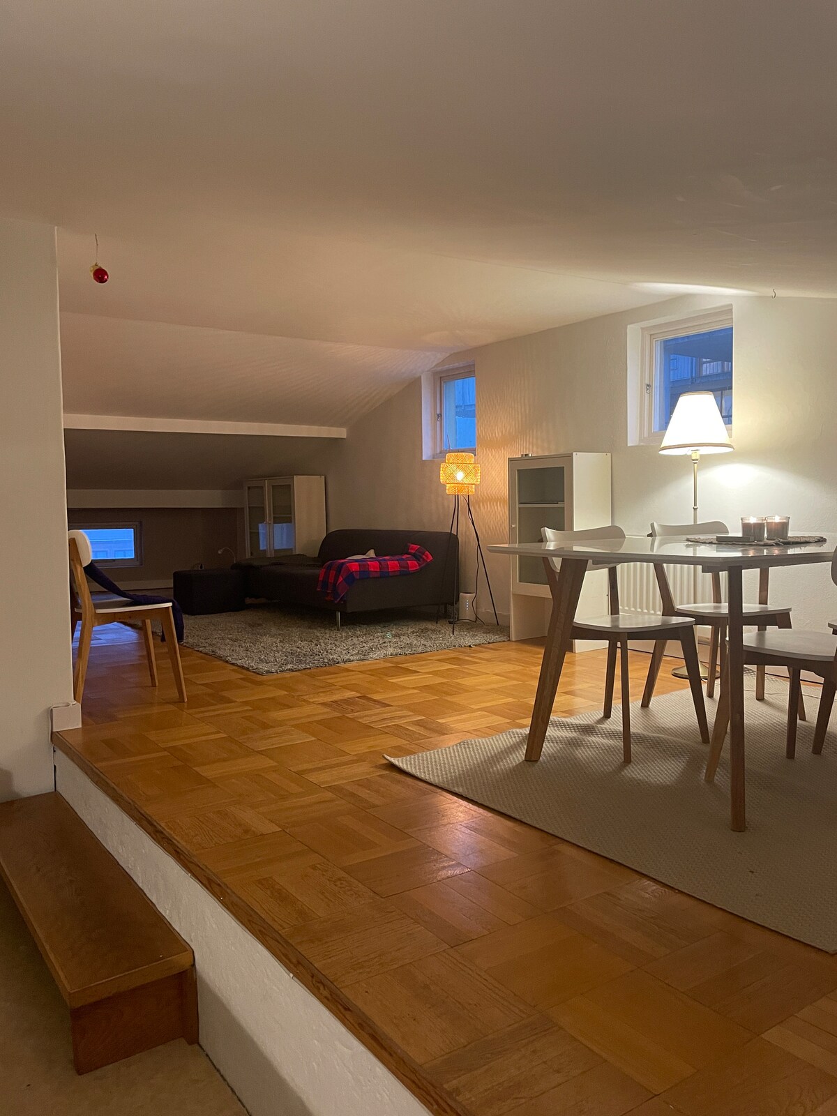 A well-lit studio space is depicted, featuring a comfortable seating area with a dark sofa draped in a red blanket. A dining table with chairs is positioned nearby, complemented by soft lighting provided by a floor lamp and a table lamp. Windows allow natural light to illuminate the room.