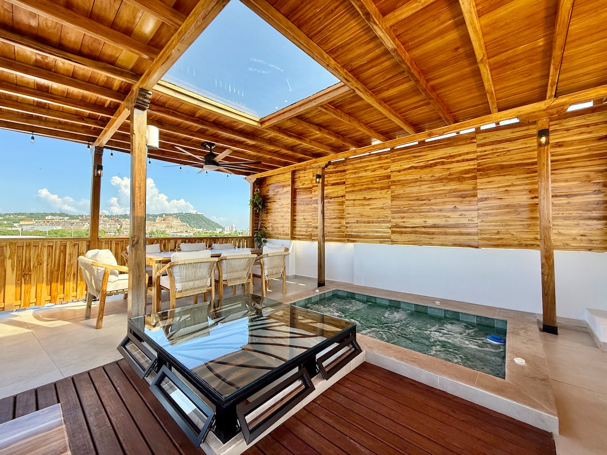 A spacious rooftop terrace features wooden ceilings and walls, providing a warm ambiance. A glass-top table surrounded by chairs sits beside a private jacuzzi. The area is elevated, offering views of the surrounding landscape through the open space above.