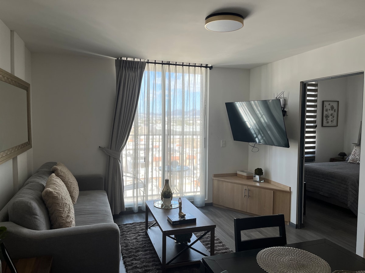 A modern living area is shown, featuring a comfortable gray sofa and an elegant coffee table. Floor-to-ceiling curtains allow natural light to flow in. A television is mounted on the wall. A doorway leads to a bedroom, while a dining area is visible in the background.
