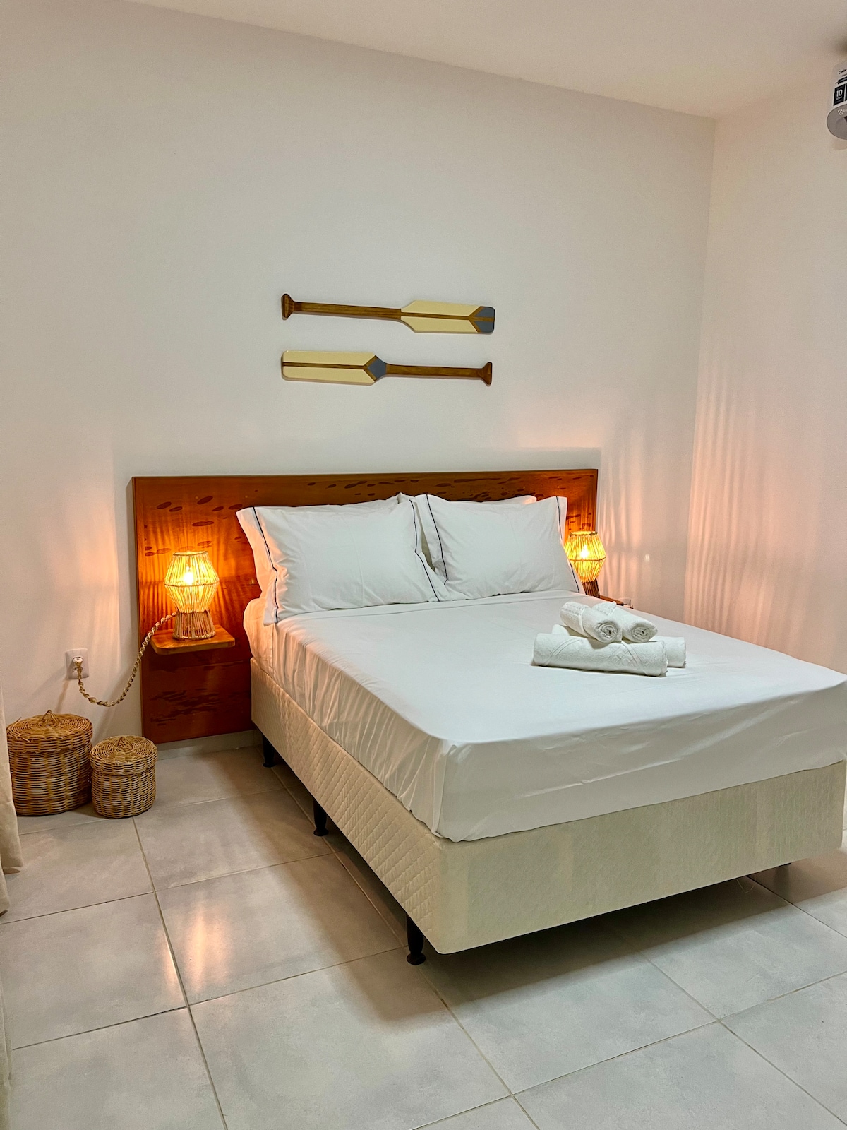A cozy sleeping area features a spacious bed with fresh linens. Two decorative oars hang on the wall above, accompanied by softly glowing bedside lamps. Woven baskets are positioned beside the bed, and the room's walls are painted in a light color, enhancing the sense of openness.