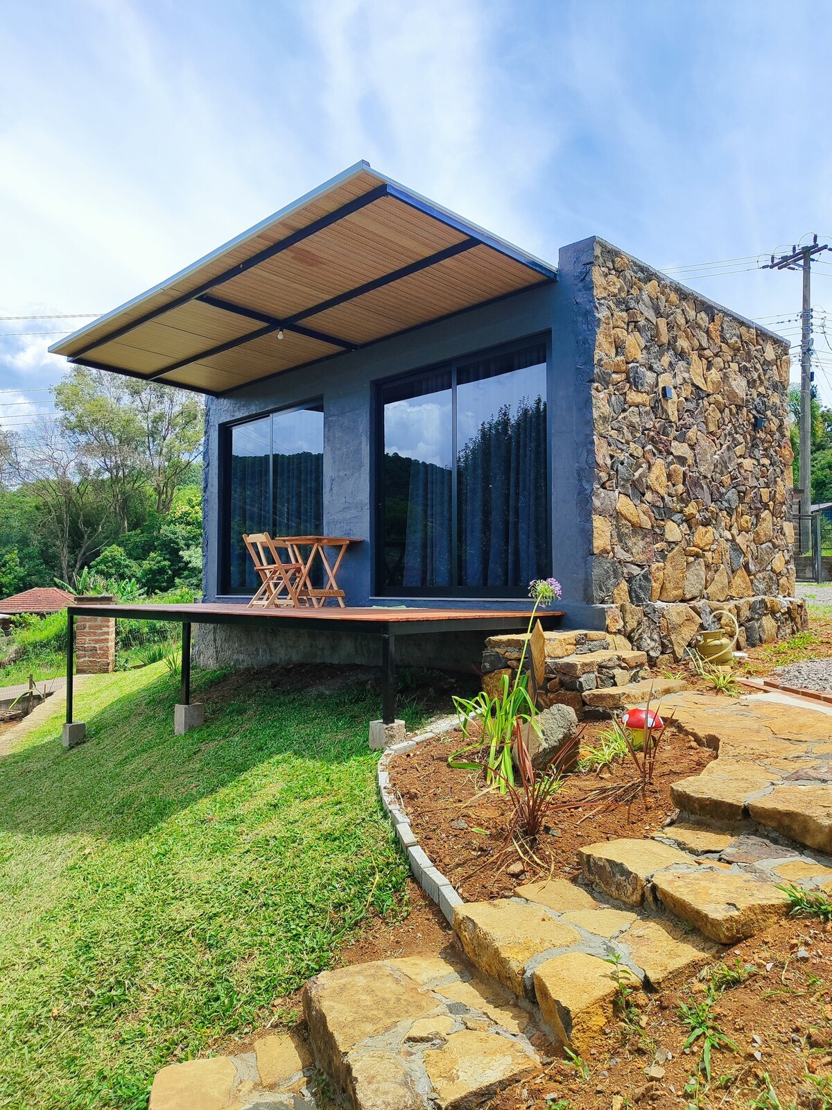 A modern cabin is shown with a stone exterior and large glass doors that open onto a deck. Two wooden chairs are positioned on the deck, overlooking a grassy area. A stone pathway leads up to the entrance, surrounded by greenery.